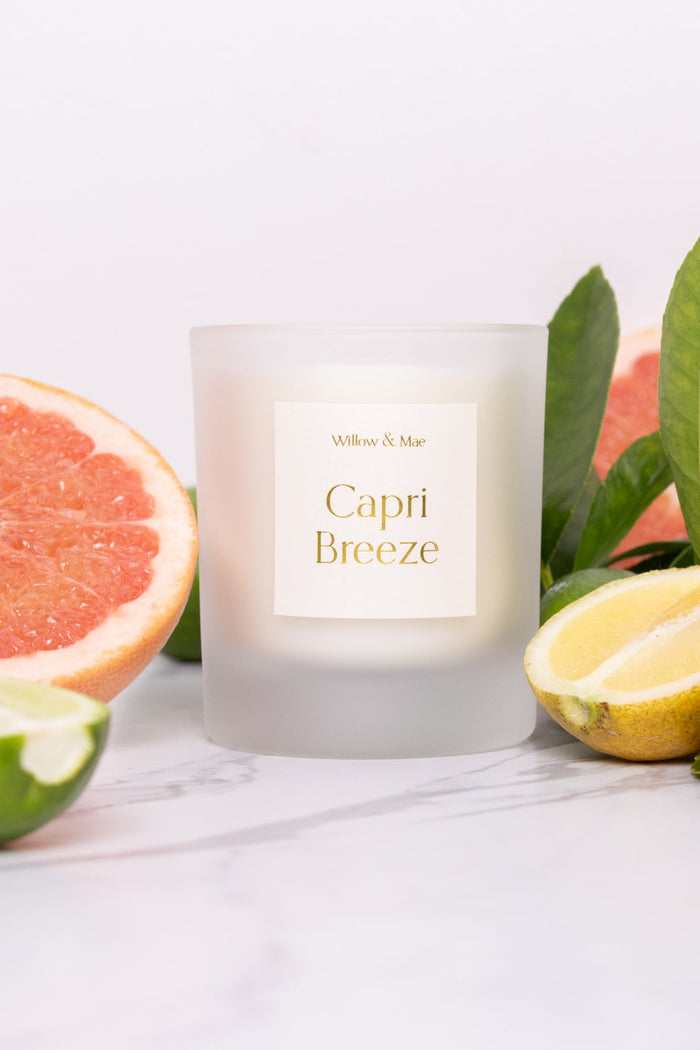 Candle labeled 'Capri Breeze' surrounded by citrus fruits and leaves on a white background