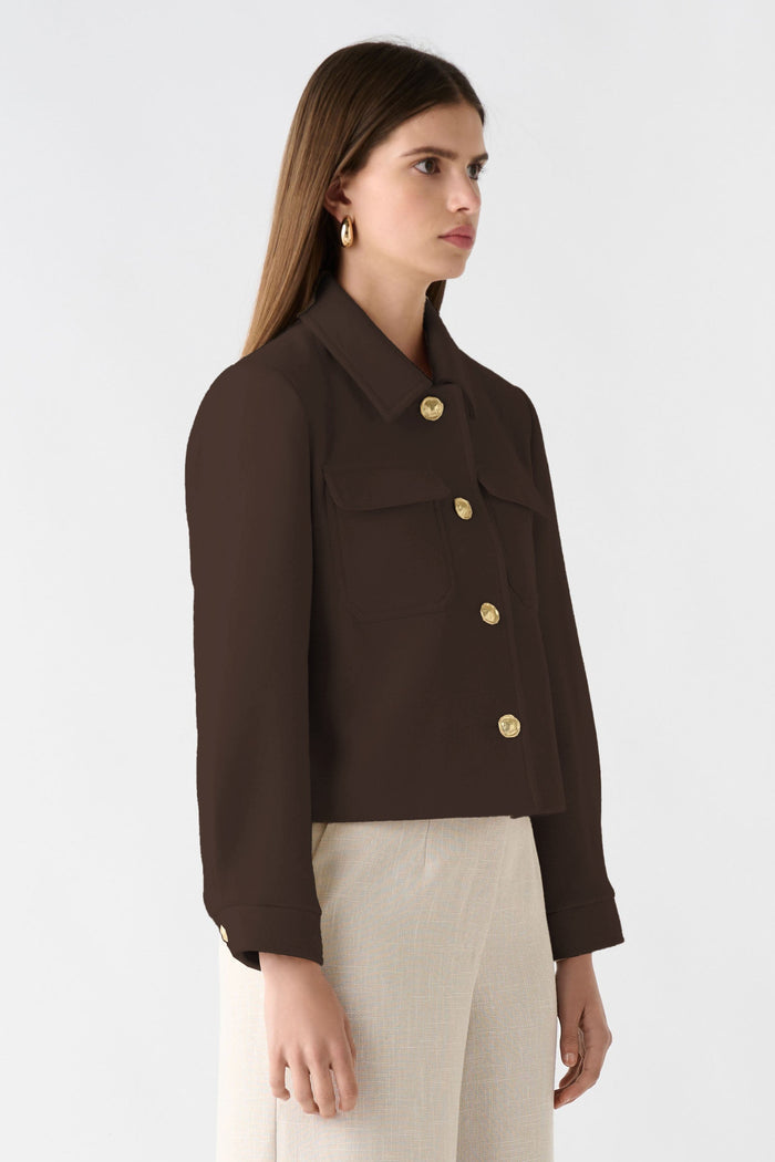 Woman wearing a brown jacket with gold buttons and beige pants on a white background