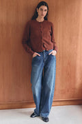 Person wearing a brown cardigan and blue jeans standing against a wooden wall.