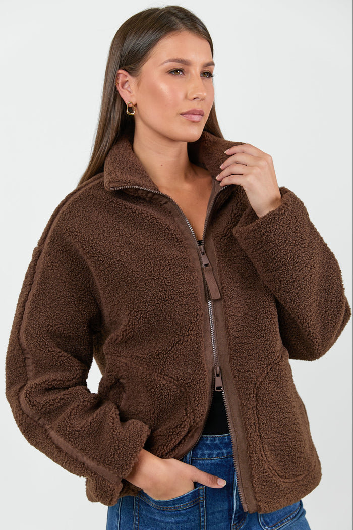 Woman wearing a chocolate brown sherpa fleece zip jacket and blue jean against a white background