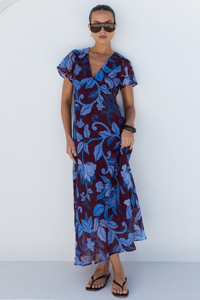 A woman modeling a short-sleeve wrap maxi dress with a blue and purple floral pattern, standing against a minimalist white wall with a circular arch.