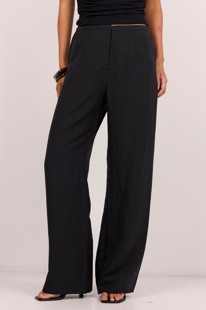 A model wearing black wide-leg trousers with a black sleeveless top and black sandals, shown from the waist down against a plain light gray studio background.