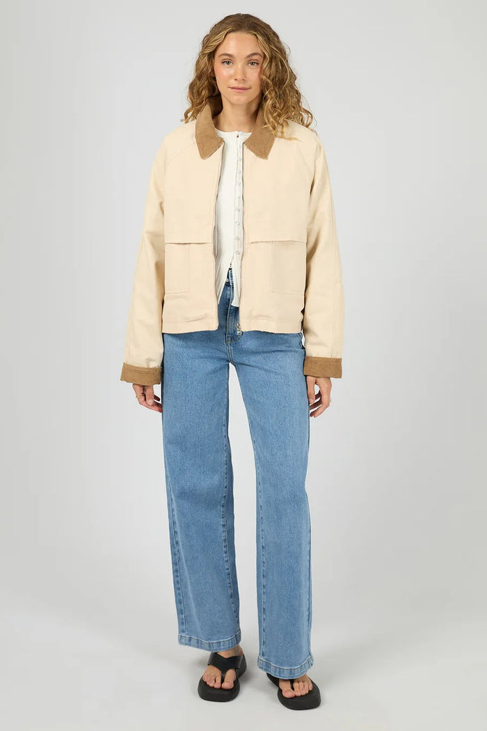 Person wearing a beige jacket with brown trim and blue jeans on a white background