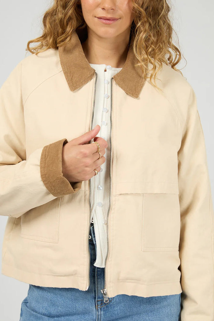 Person wearing a beige jacket with brown collar and cuffs on a plain background