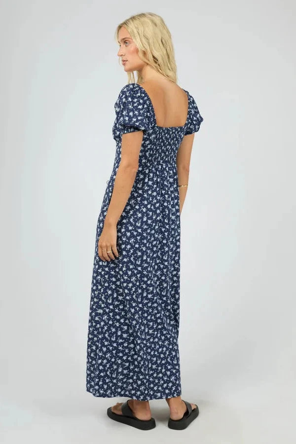 A woman modeling a navy blue maxi dress with a white floral pattern, featuring puff sleeves and a smocked bodice, shown from a three-quarters back view against a light gray studio background.
