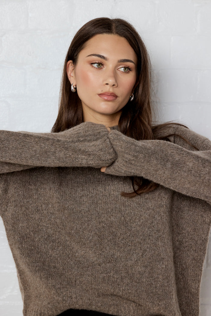 Woman wearing a brown sweater against a white brick wall.