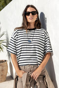 A woman wearing a black and white horizontally striped short-sleeve t-shirt, beige cargo pants, and black sunglasses, leaning against a white stucco wall outdoors.