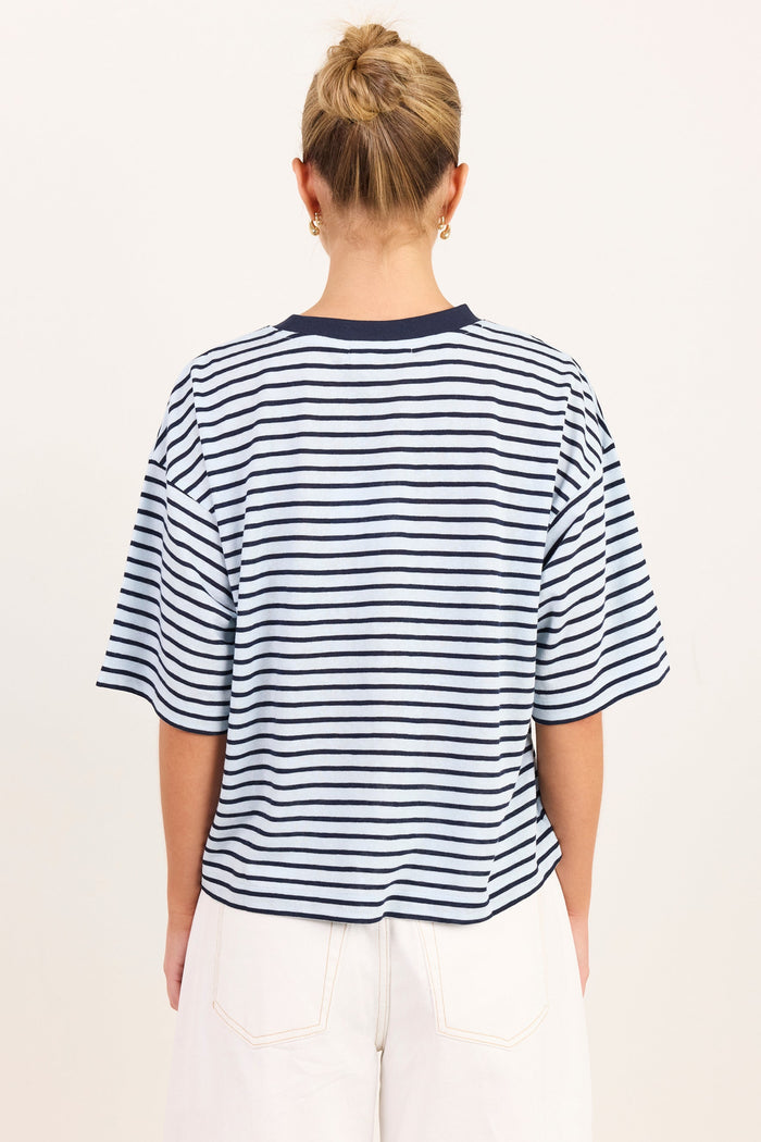 A woman viewed from behind wearing a navy and white horizontally striped short-sleeve t-shirt and white pants, set against a plain off-white studio background.