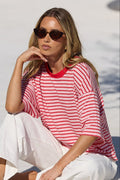 A woman wearing a red and pink striped t-shirt and white pants sits on the edge of a swimming pool, wearing sunglasses.
