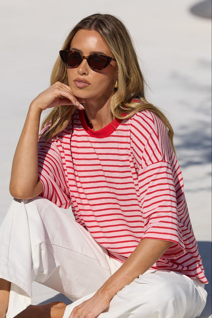 A woman wearing a red and pink striped t-shirt and white pants sits on the edge of a swimming pool, wearing sunglasses.
