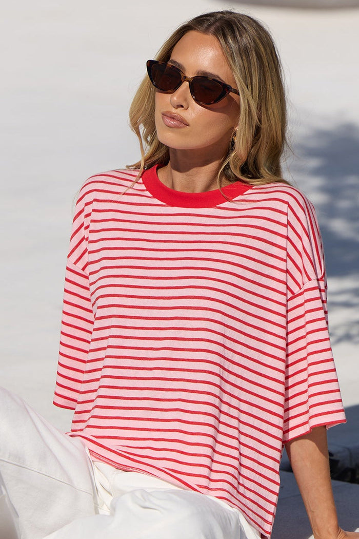 A woman wearing a red and white striped t-shirt and white pants sits on a white tiled pool deck next to a tree.