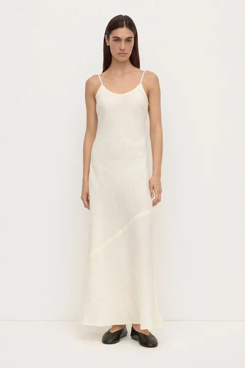 A woman modeling a long, sleeveless white slip dress with thin spaghetti straps and black flat shoes, standing against a plain white studio background.