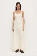 A woman modeling a long, sleeveless white slip dress with thin spaghetti straps and black flat shoes, standing against a plain white studio background.