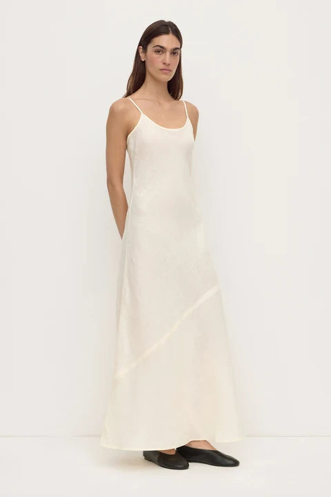 A woman modeling a long, sleeveless cream-colored slip dress with thin straps and black flat shoes, standing against a plain white studio background.