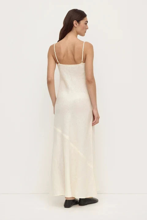 A woman shown from the back wearing a long, sleeveless cream slip dress with thin straps and black shoes, standing against a plain white studio background.