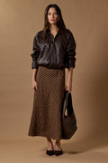 Woman wearing a brown leather jacket and patterned skirt holding a matching handbag against a brown background