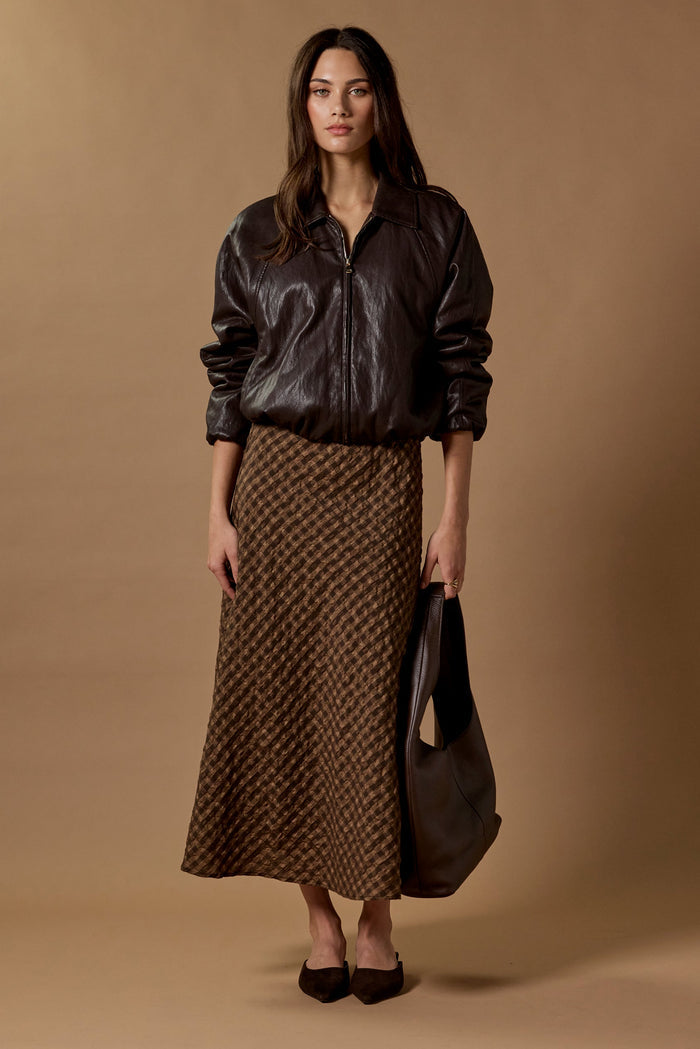 Woman wearing a brown leather jacket and patterned skirt holding a matching handbag against a brown background