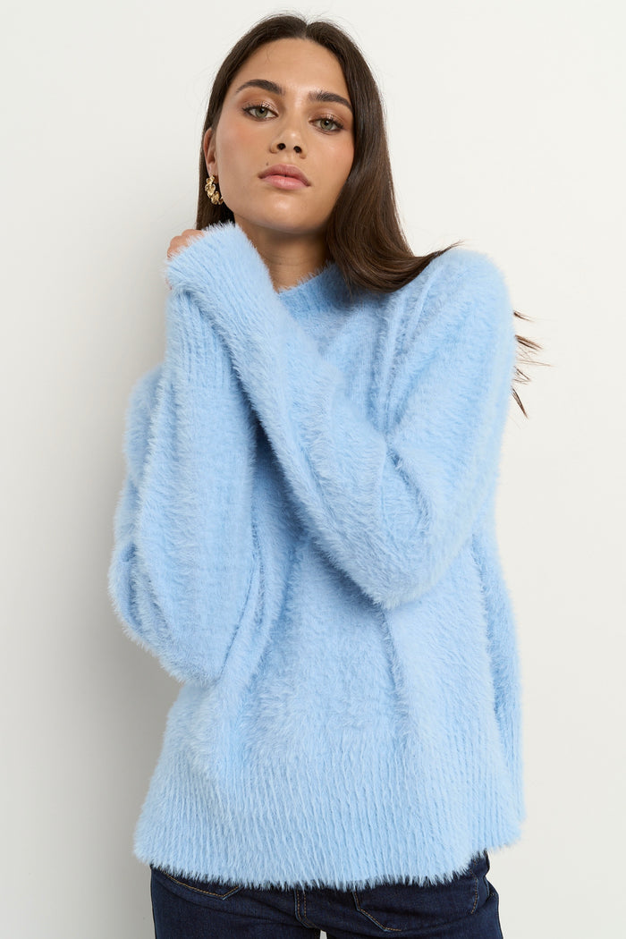 Woman wearing a light blue fuzzy funnel neck knit with relaxed fit and long sleeves, paired with dark wide-leg jeans.