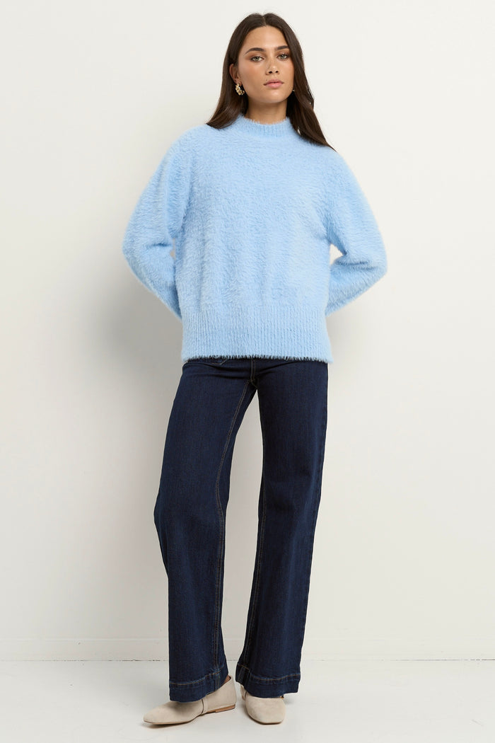 Woman wearing a light blue fuzzy funnel neck knit with relaxed fit and long sleeves, paired with dark wide-leg jeans.