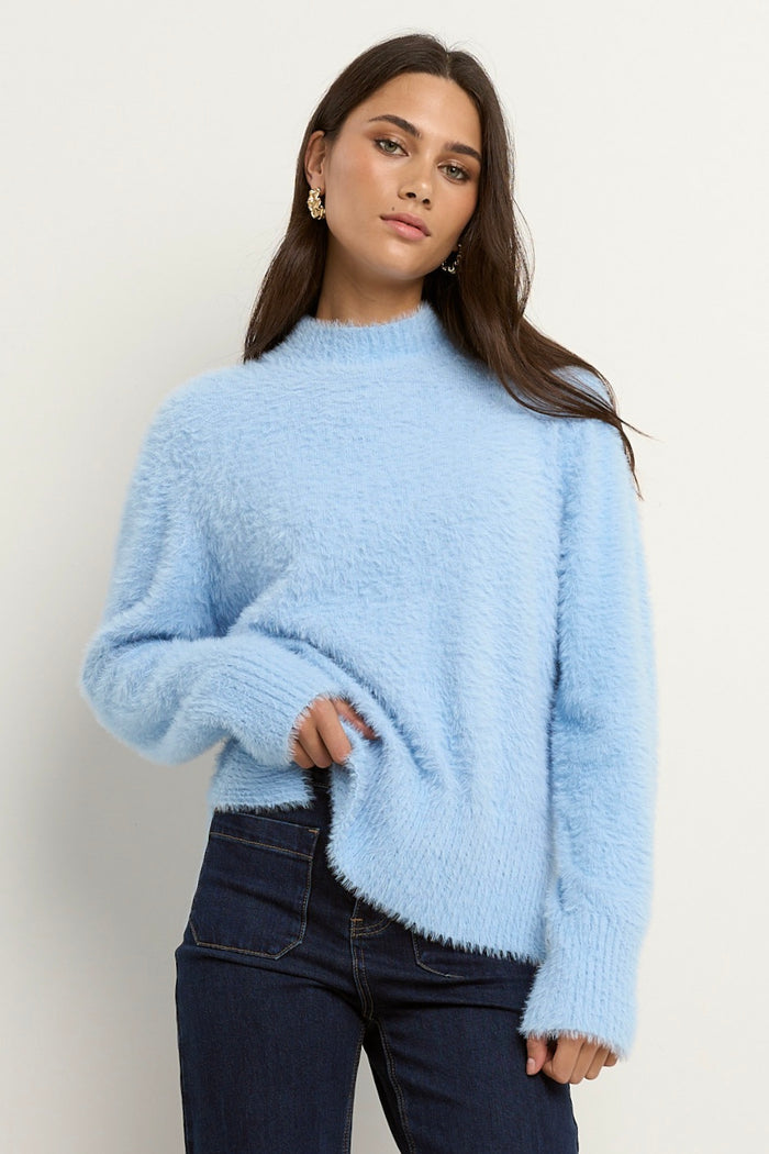 Woman wearing a light blue fuzzy funnel neck knit with relaxed fit and long sleeves, paired with dark wide-leg jeans.