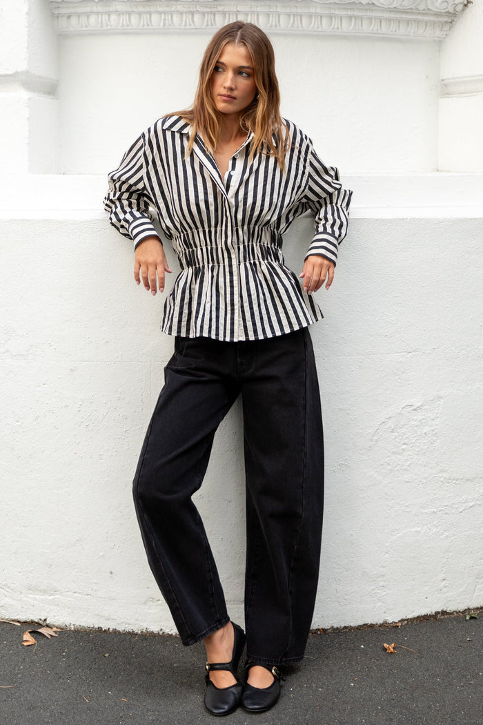 Woman in a striped shirt and black pants standing against a white wall.