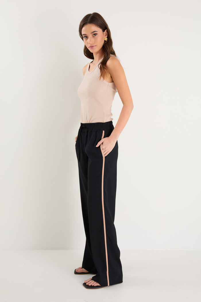 Woman wearing a beige sleeveless top and black pants with a gold stripe on a white background