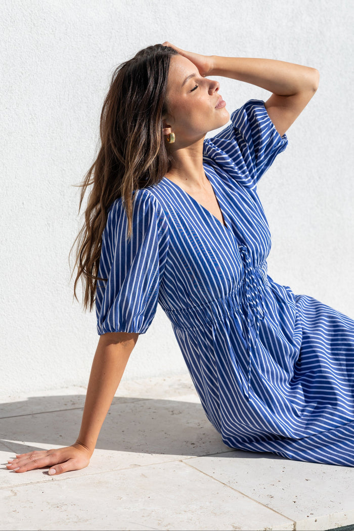 A woman wearing a blue and white vertically striped midi dress with puff sleeves and a gathered waist, posing on a light stone surface against a white textured wall.