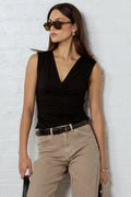 A woman stands against a white brick wall wearing a black sleeveless wrap top, beige cropped pants, a black belt, black-and-brown patterned flat shoes, sunglasses, and holding a black clutch and a beige duffel bag.
