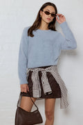 Woman wearing a light blue sweater and striped skirt with a brown handbag against a white wall.