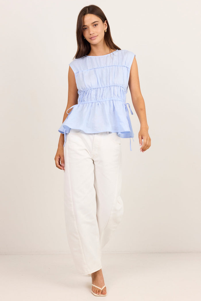 A woman modeling a light blue sleeveless peplum top paired with white wide-leg pants and white sandals, standing against a plain off-white studio background.