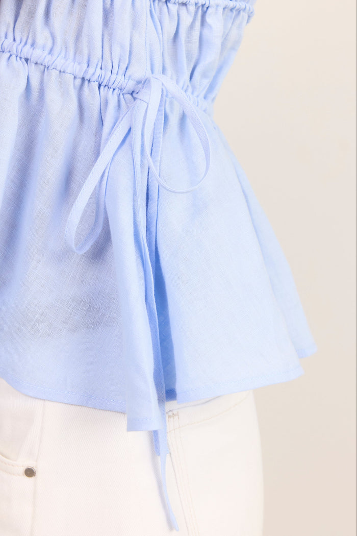 Close-up of a light blue peplum-style top with an elasticized gathered waist and a side tie, worn with white pants against a plain light beige background.