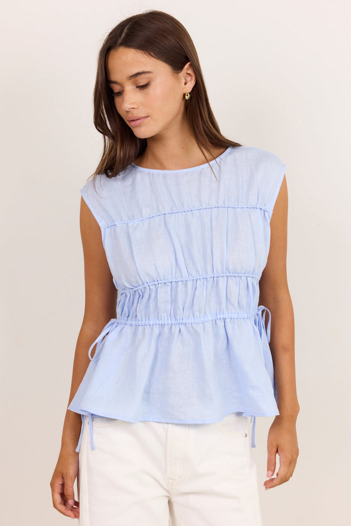 A woman wearing a light blue sleeveless peplum top with a smocked waist and white pants, standing against a plain off-white background.