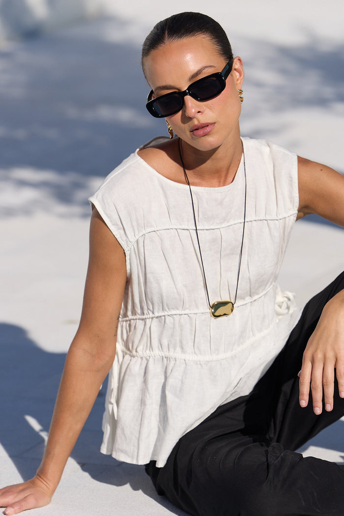 A woman wearing a white sleeveless peplum top, black pants, and black sunglasses sits on a concrete poolside ledge in bright sunlight.