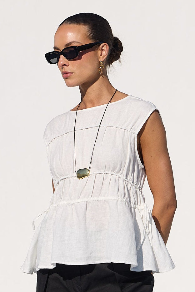 A woman stands by a pool wearing a white sleeveless peplum top, black cropped wide-leg pants, and black sandals in a bright white architectural setting.