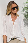 A woman wearing a white three-quarter sleeve button-up blouse and a beige mini skirt, styled with black sunglasses and a black belt with a gold buckle, standing against a white wall with a green plant in the background.