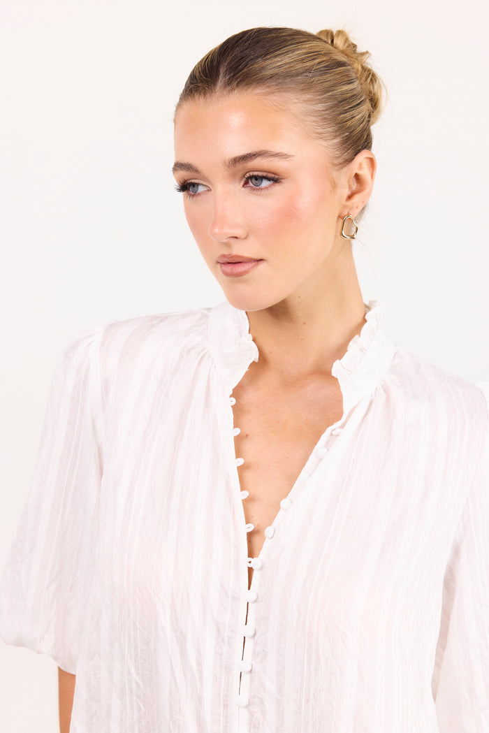 A woman with blonde hair in a bun wearing a white crinkled blouse with a ruffled collar and button-front closure, photographed against a plain white background.