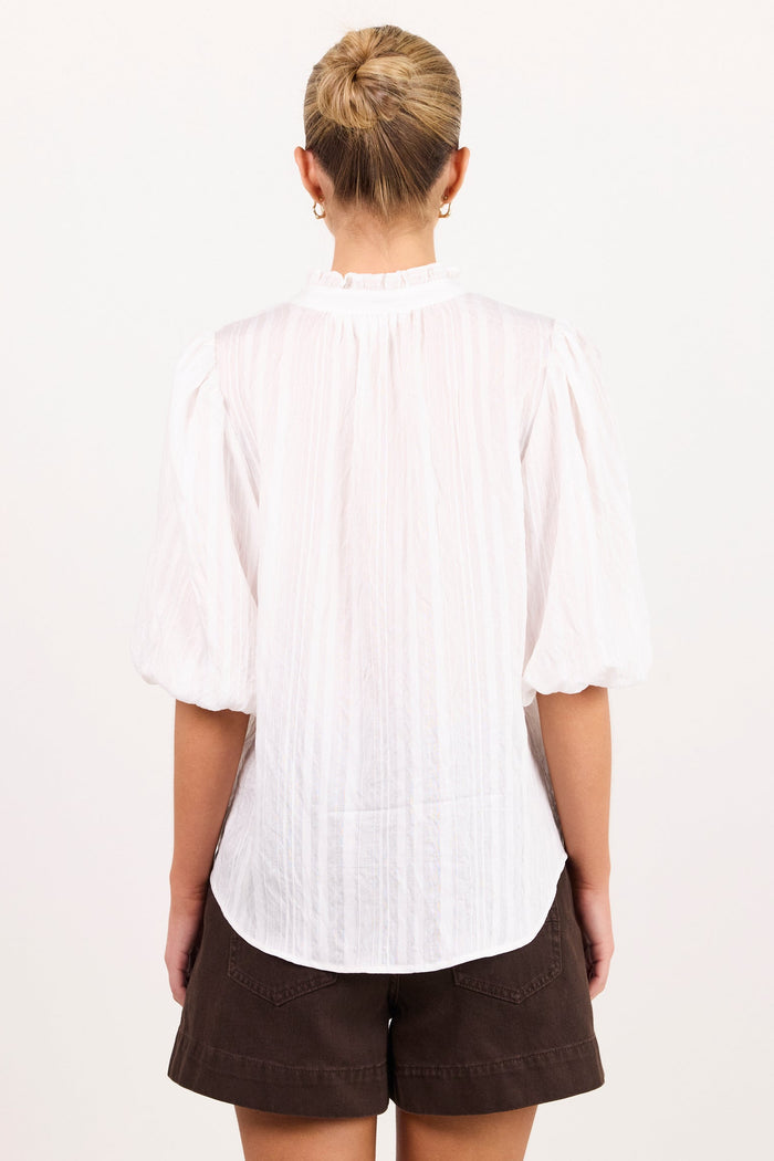 A woman viewed from behind wearing a white pleated blouse with puff sleeves and dark brown shorts, standing against a plain white background.
col_pattern