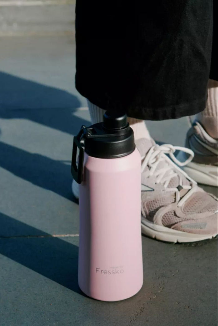 Pink water bottle with black lid on a concrete surface, person wearing black pants and white sneakers in the background.