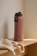 A dusty rose pink Fresko water bottle with a dark gray lid and carrying loop, standing on a light wood bench next to a beige woven bag.