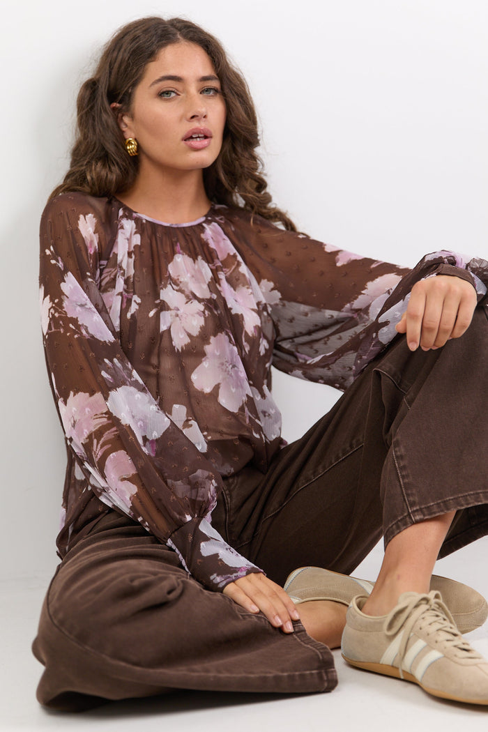 Woman wearing a floral blouse and brown pants sitting on the floor against a white background