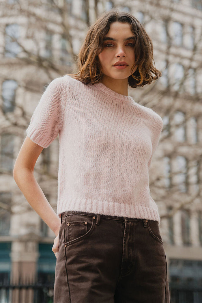 Woman wearing a light pink sweater and dark pants standing in front of an architectural building.