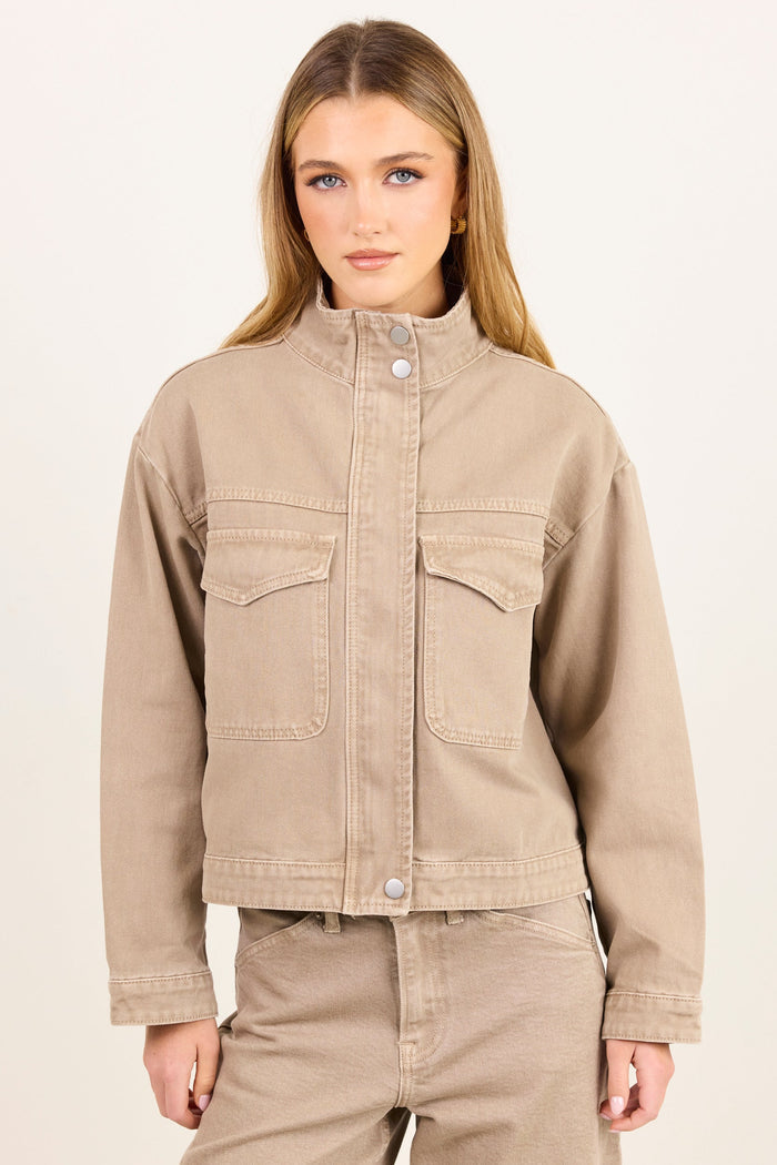 A woman modeling a cropped beige denim jacket with a stand-up collar and front flap pockets, paired with matching beige pants, against a plain light background.