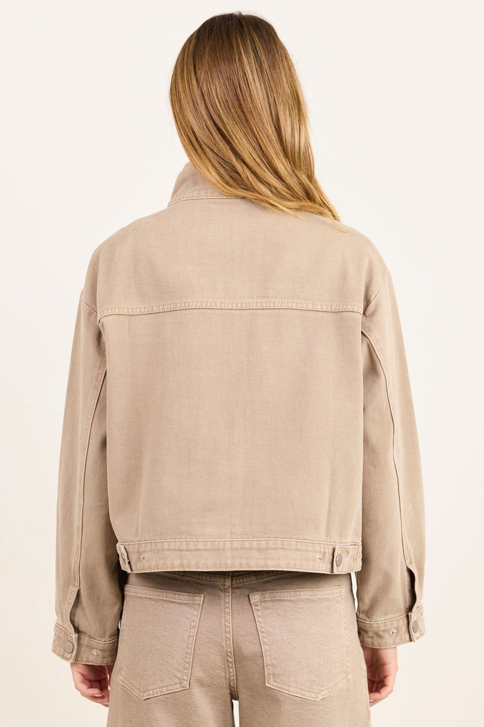 A woman viewed from behind wearing a cropped beige denim jacket and matching beige pants against a plain off-white studio background.