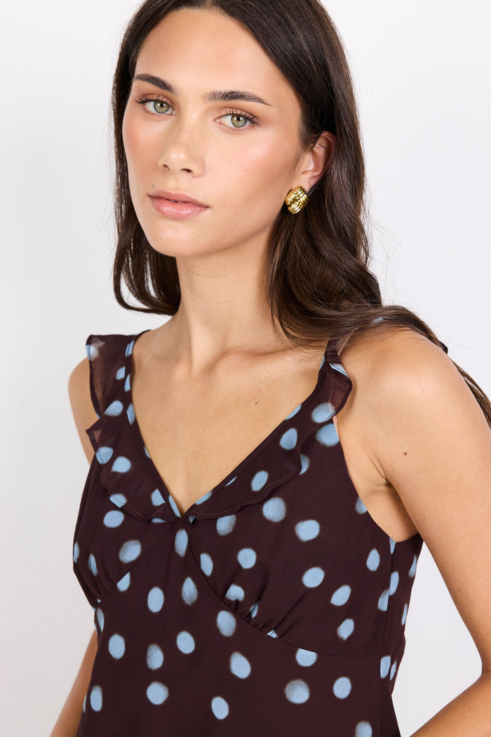 A woman wearing a dark brown sleeveless top with light blue polka dots and ruffled shoulder details, paired with a gold geometric stud earring, against a plain white background.