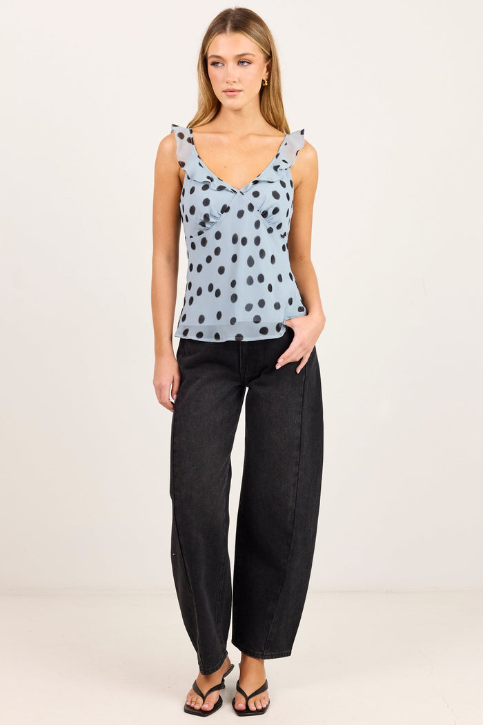 A woman modeling a sleeveless light blue top with black polka dots and black wide-leg pants against a plain off-white studio background.