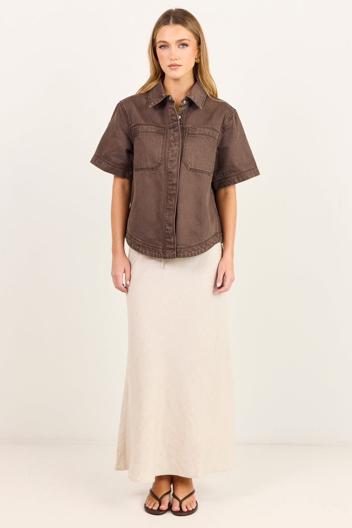 A woman stands against a plain white studio background wearing a short-sleeve brown button-up shirt and a long cream maxi skirt.