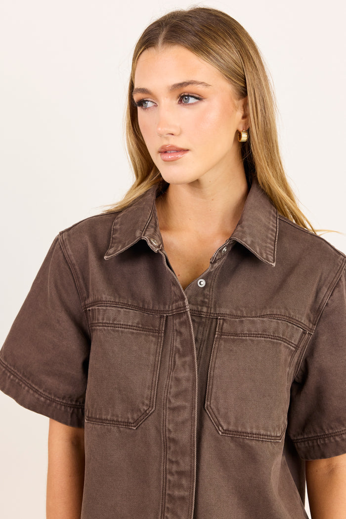 A woman wearing a brown short-sleeve button-up shirt with chest pockets, photographed against a plain light background.