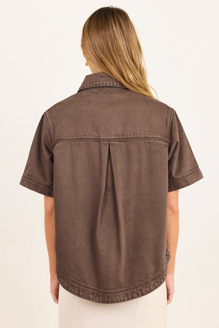 A woman viewed from the back wearing a brown short-sleeve button-up shirt with a central back pleat, paired with a light cream skirt, against a plain off-white background.