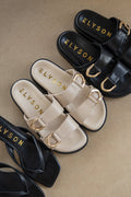 Elyson sandals in black, beige, and gold on a beige surface