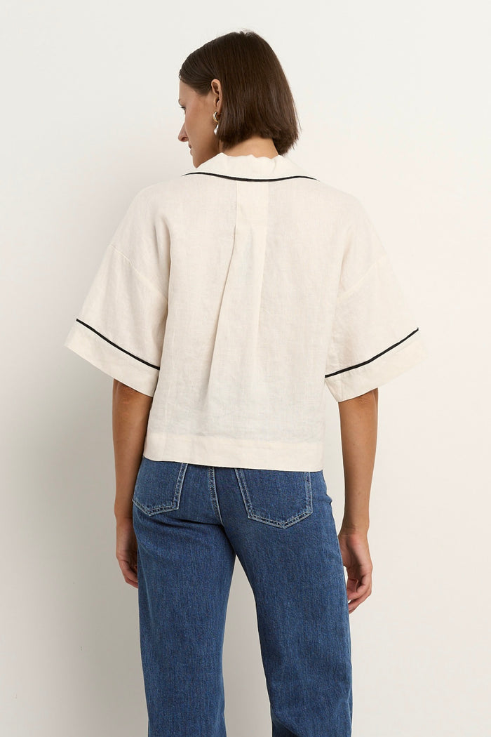 Woman wearing an oversized cream linen shirt with black piping, short sleeves, and front buttons, styled with blue denim jeans.